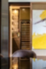Sliding screw door with framed picture. Wine fridge with glass shelves filled with bottles and lit glasses, beside colorful abstract painting. A cozy, elegant setting. MAD Studio