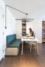 Woman organizing shelves in a modern dining area with teal seating, a white table, and dark wood floors. Stylish geometric light overhead. MAD Studio