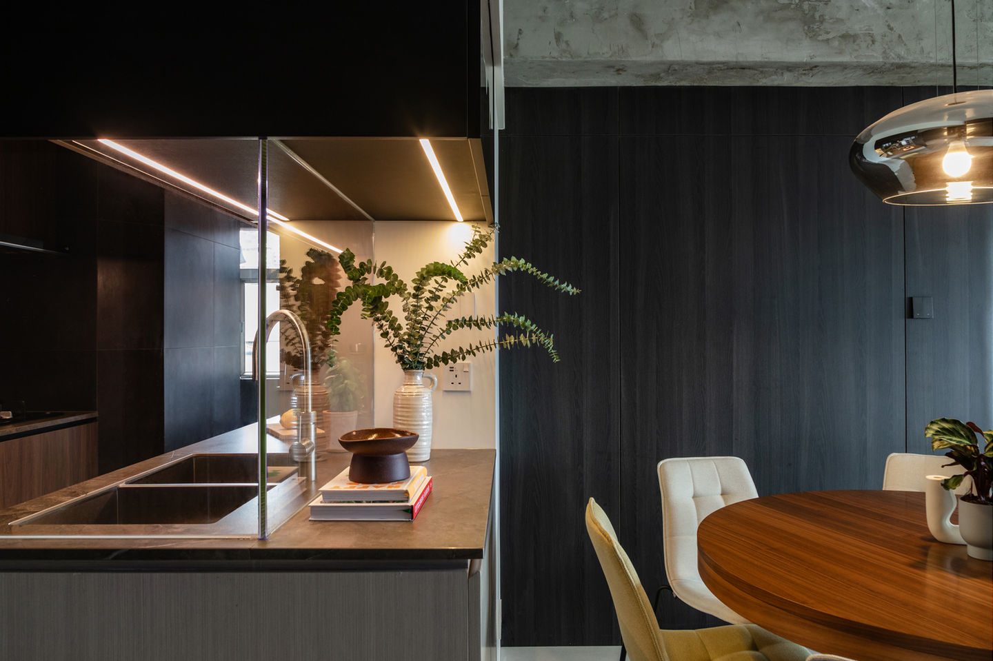 Modern kitchen and dining area featuring a sleek dark wood wall, ambient lighting, and an elegant round wooden table. A chic countertop with a stainless steel sink, a stylish white vase with green foliage, and contemporary decor highlight the luxury interior design. Ideal for showcasing minimalist aesthetics and sophisticated living spaces. 