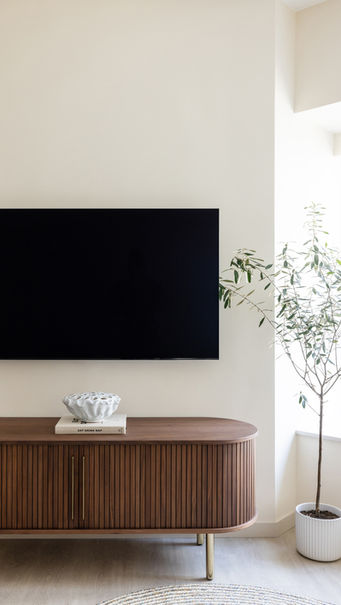 A modern living room featuring a sleek, mounted flat-screen TV above a stylish wooden console with vertical slats and gold handles. The console is adorned with a decorative bowl on top of a coffee table book. To the right, a potted plant adds a touch of greenery near a large window, complementing the minimalist and elegant design, Island Harbourview, Residential interior design
