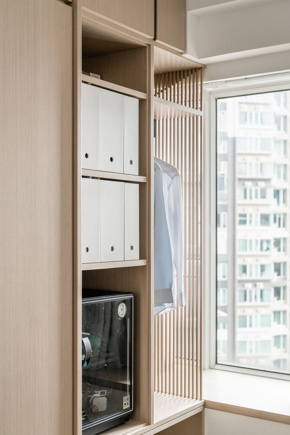 Modern interior design featuring custom built-in wooden shelving with sleek storage, showcasing minimalist home organization. The shelving includes a mix of open and closed spaces, complemented by a stylish wardrobe area and large window offering natural light. Ideal for urban residential interiors and smart storage solutions.