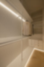 Modern walk-in closet with illuminated shelves. Beige shirts hang neatly. Light wood floor, white drawers, minimalistic design.