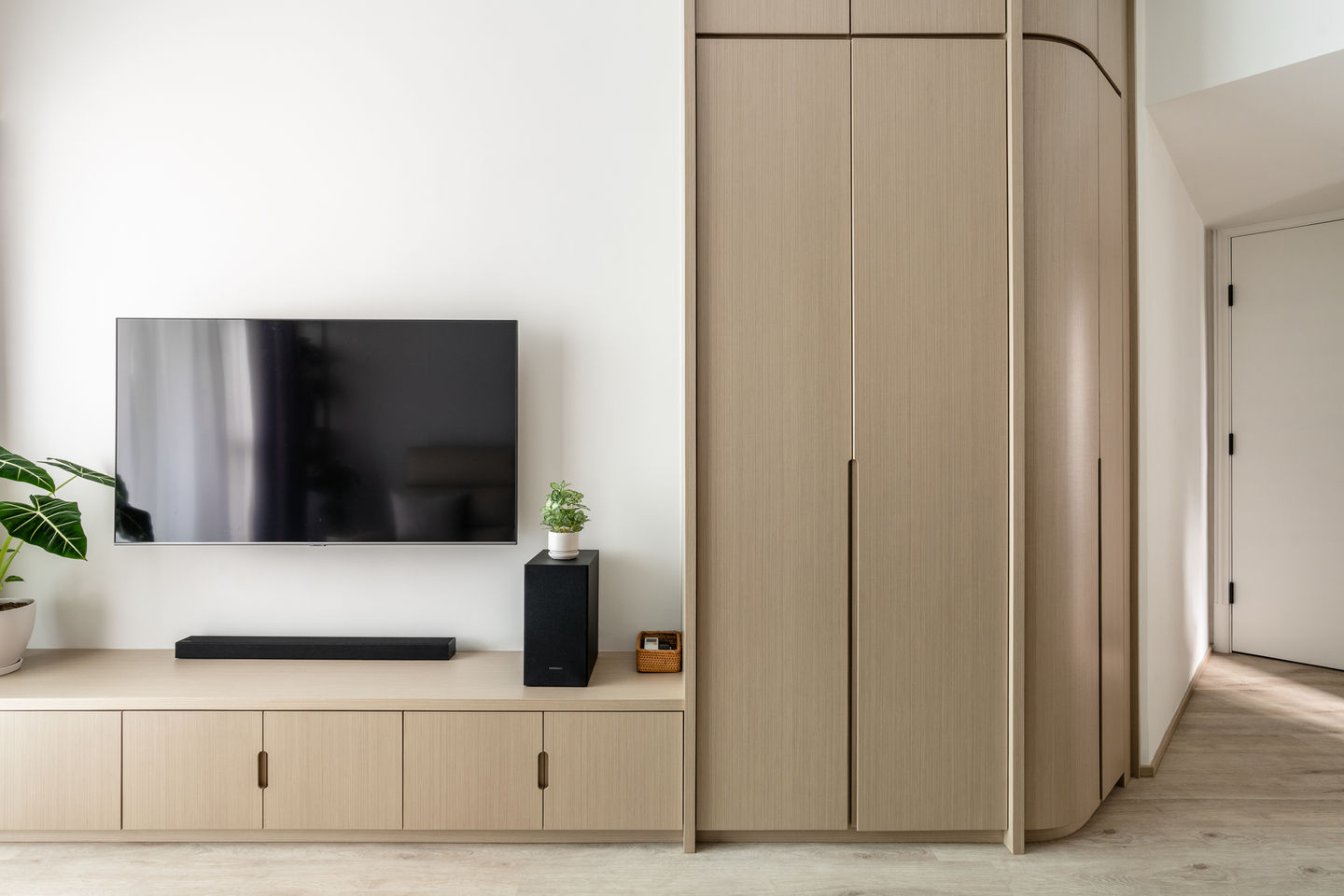 Modern minimalist living room interior design featuring a sleek wall-mounted flat-screen TV, light wood cabinetry, and built-in storage. A potted plant adds a touch of greenery, enhancing the clean and contemporary aesthetic. Perfect for small spaces, this stylish setup exemplifies functional and elegant design. 