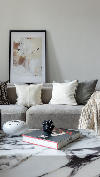 A modern living room designed by MAD Studio featuring a stylish gray sectional sofa adorned with assorted neutral-colored cushions. A textured, cream-colored throw is draped elegantly over the arm of the sofa. Above, an abstract art piece in a black frame adds visual interest to the minimalist beige wall. A chic marble coffee table in the foreground holds a designer book, modern decorative objects, and a ceramic piece, enhancing the sophisticated ambiance. Ideal for showcasing contemporary interior design inspiration.
