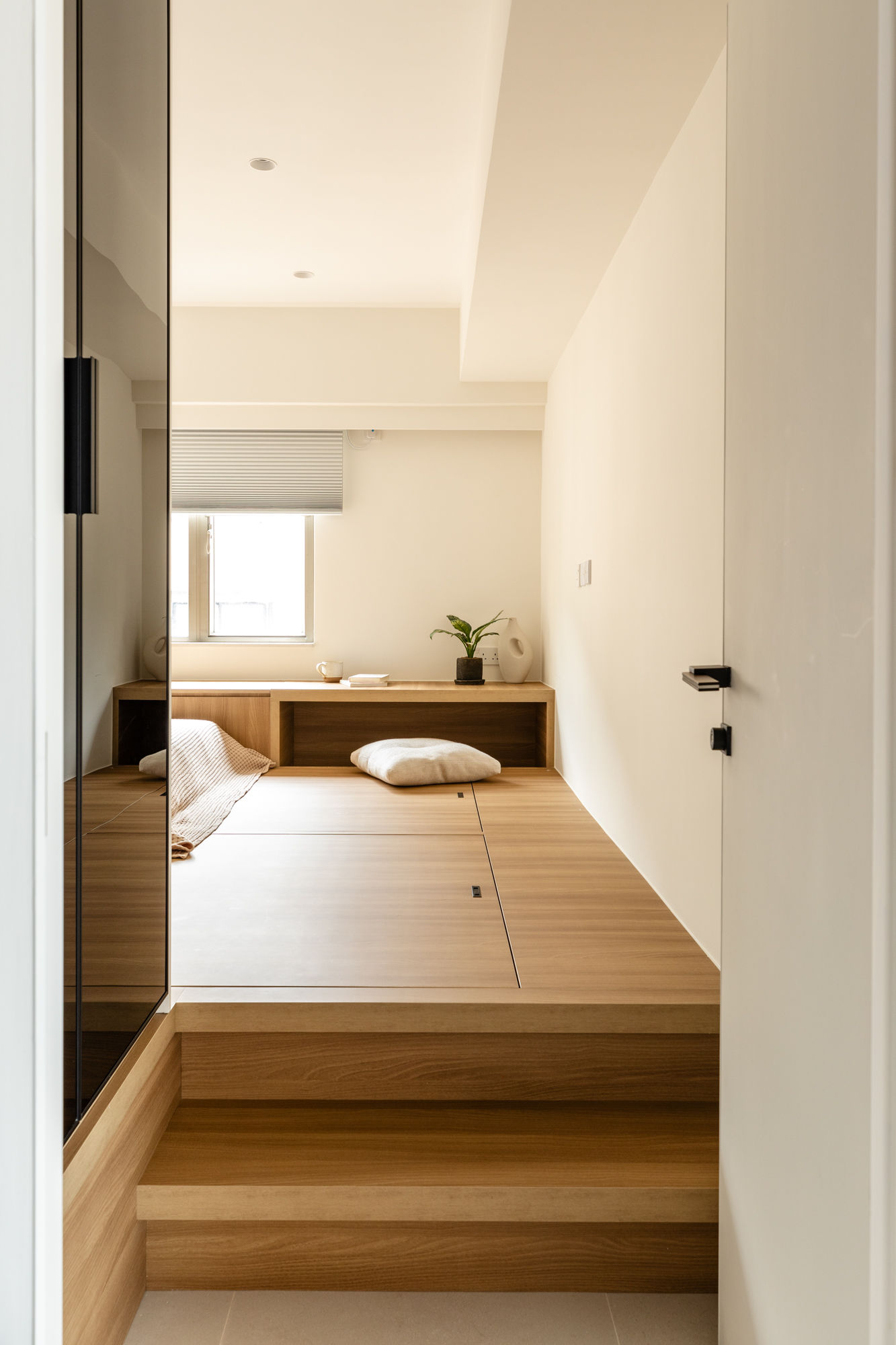 Minimalist Japanese-inspired bedroom design featuring a raised wooden platform bed with built-in storage. The room is bathed in natural light from a window with a neutral roller shade, enhancing the calm and serene interior.