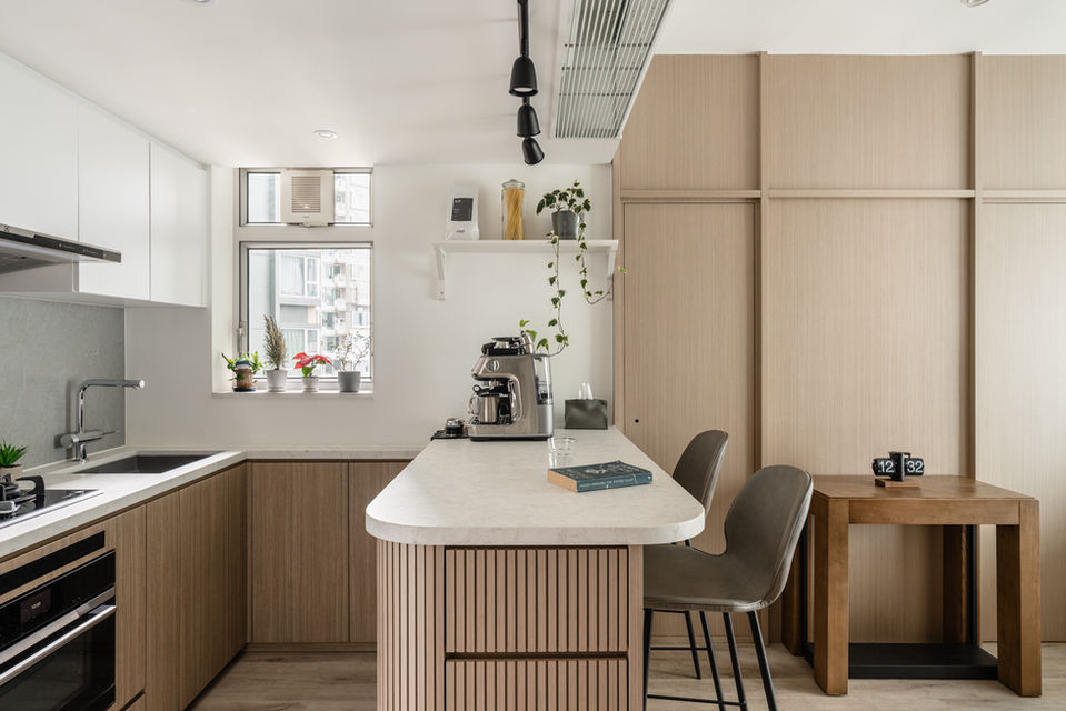 Modern kitchen interior design featuring a minimalist aesthetic, with sleek wood cabinetry and a central island topped with light quartz countertops. The space is accented by stylish bar stools, stainless steel appliances, a built-in cooktop, and subtle decorative plants on the windowsill. Ideal for showcasing contemporary interior design trends and maximizing small kitchen spaces. Perfect for interior design inspiration and home renovation ideas.