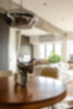 Modern living room interior featuring a stylish round wooden dining table with a decorative plant centerpiece. The open-concept space includes a comfortable beige sofa, large windows for natural light, and contemporary design elements like a metallic pendant light and exposed concrete beams. Ideal for showcasing luxury interior design, home decor inspiration, and urban apartment styling.