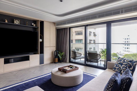 Modern living room, linear aircon grille, blue area rug with the border, beige textile sectional sofa, wood TV cabinet with black colour scheme, beige textile ottoman, balcony with rattan lounge chairs, grey curtains