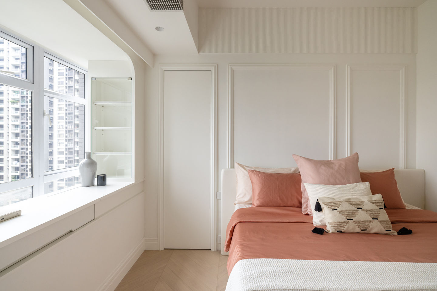 Stylish modern bedroom interior designed by MAD Studio, showcasing minimalist decor and natural lighting. Features include a cozy bed with peach and ivory pillows, sleek built-in shelving, and large windows offering city views. Ideal for contemporary home design inspiration and professional interior decorating services.